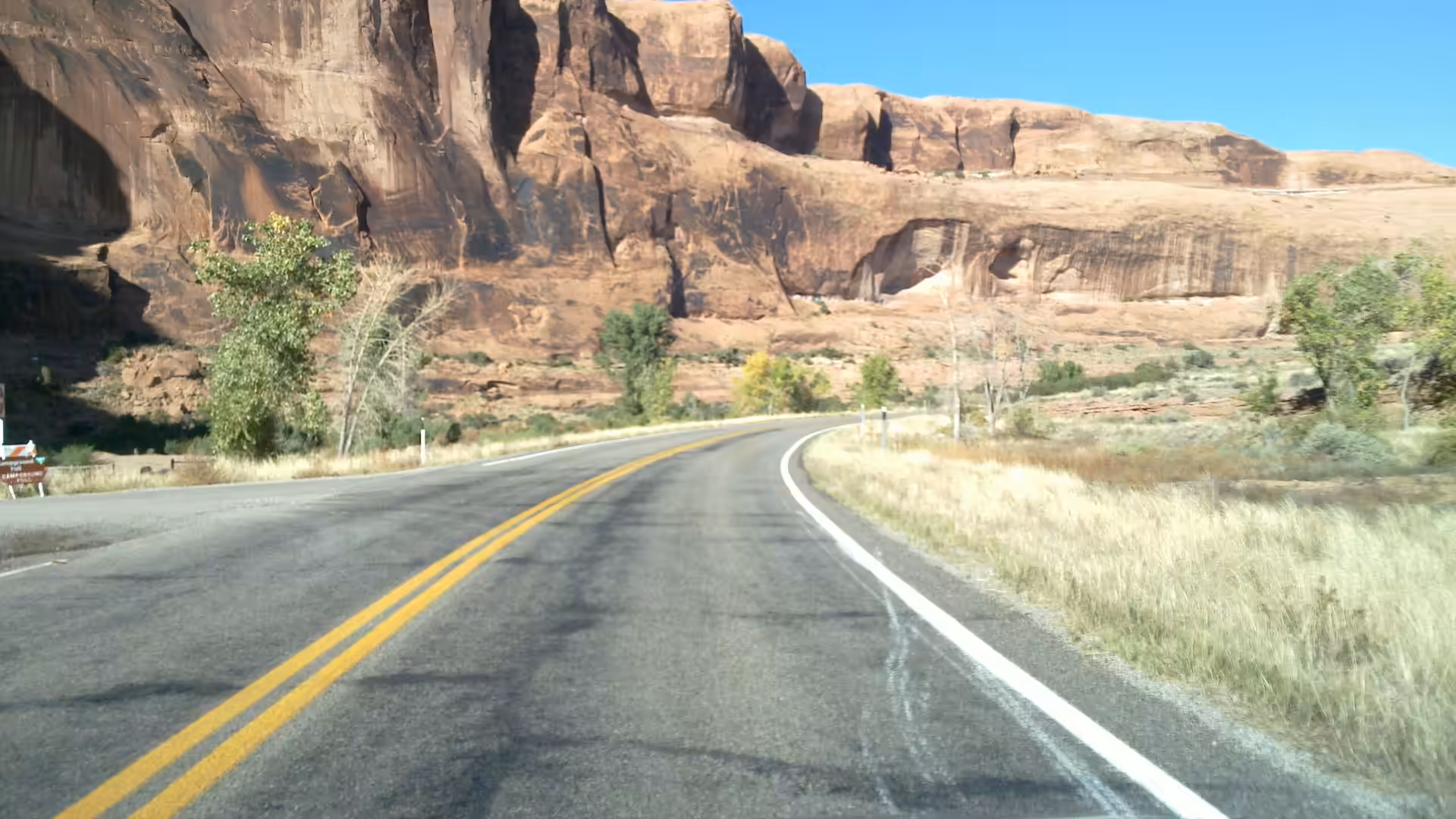 Paved desert highway between red sandstone cliffs with roadside trees.