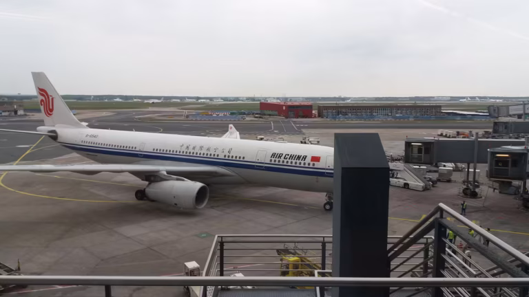 An Air China passenger aircraft parked at an airport gate, viewed through the terminal windows beneath an overcast sky.