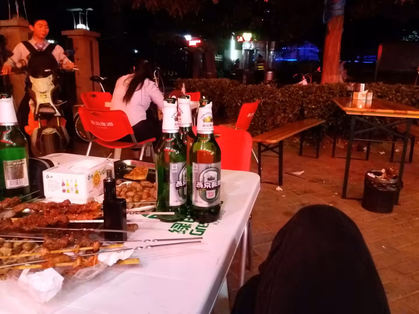 Nighttime outdoor dining scene in Beijing with grilled skewers, green beer bottles, red plastic chairs, bicycles, and other diners under warm red lighting.