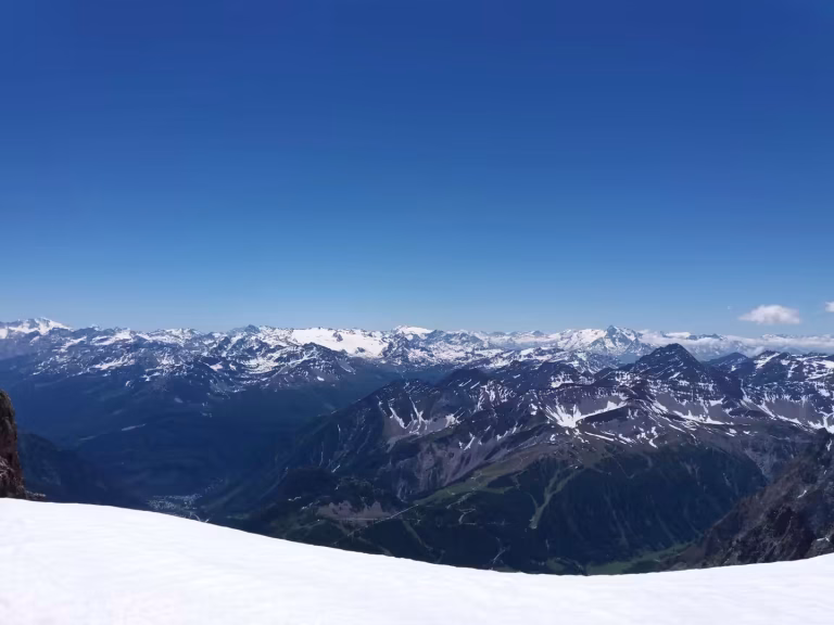 From a bright snowfield, snowy mountain ridges stretch across a wide valley beneath a blue sky.
