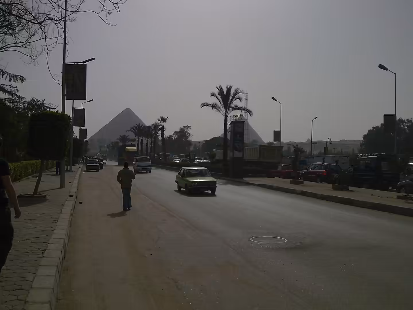 Palm lined city road with cars and three ancient pyramids visible in bright dusty distance.