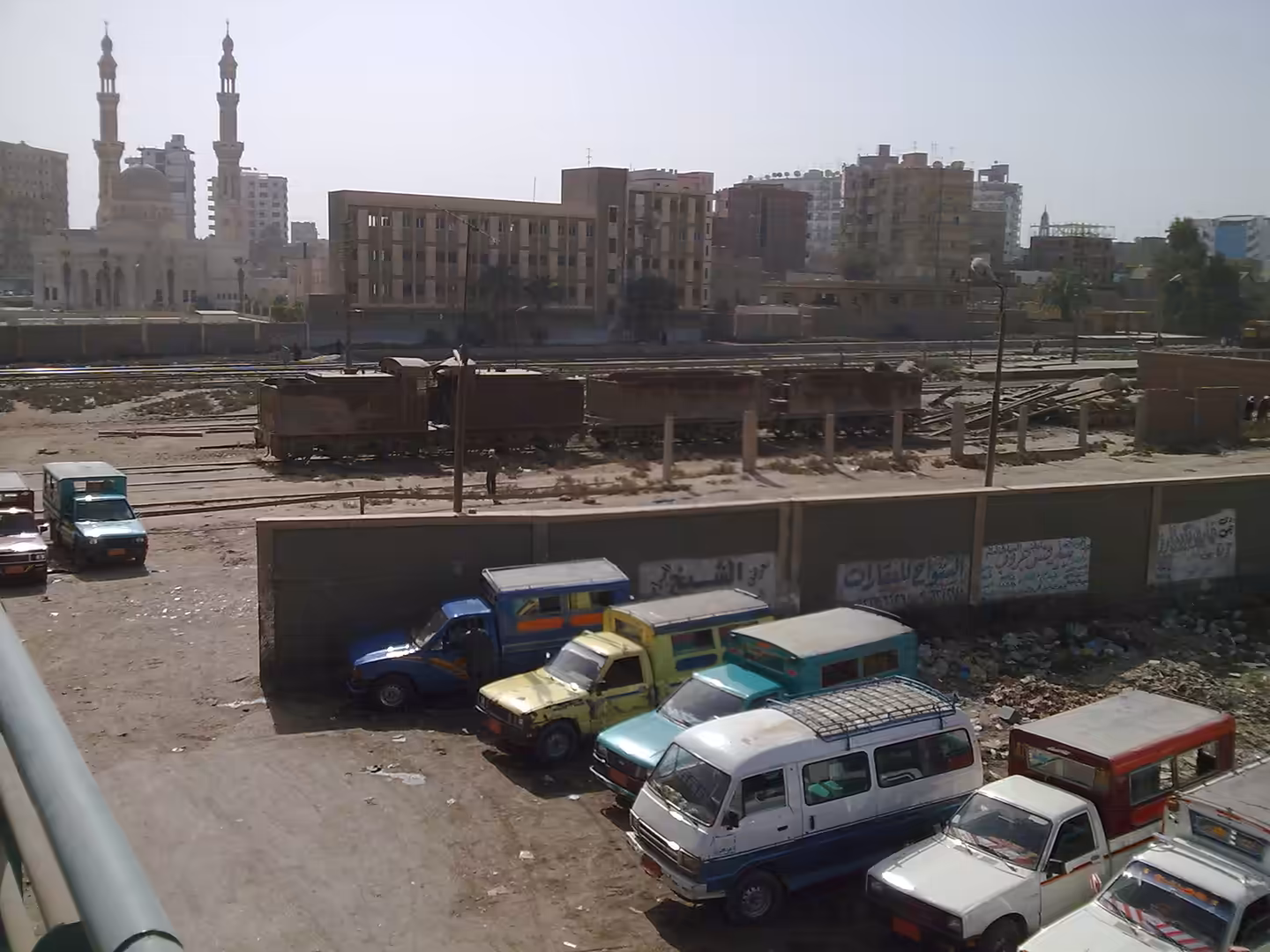 Mosque minarets above rail yard with rusted tank cars, dusty lot, and parked vans in dense city area.