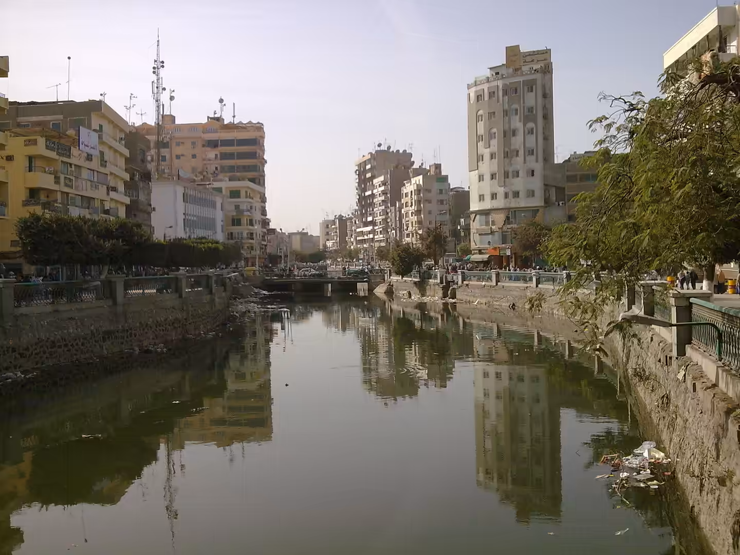 City canal with still water reflections, bridge, apartments, and tree lined embankments.