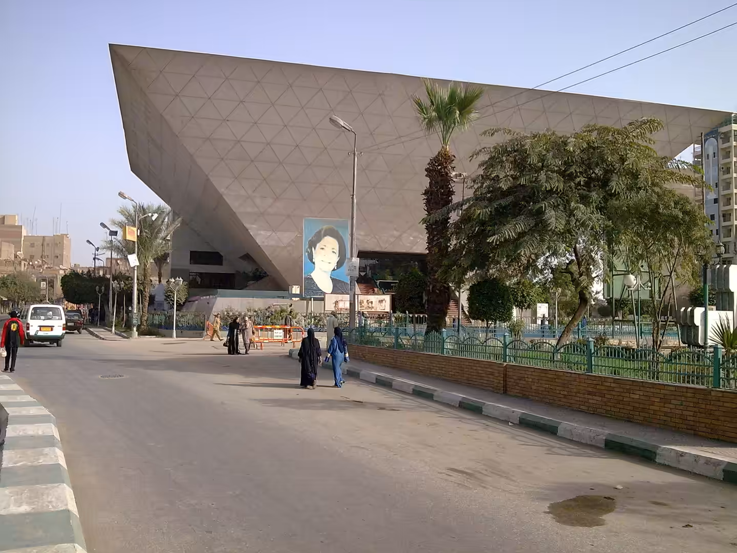 Angular modern building with geometric facade, palm trees, street pedestrians, and large portrait banner.
