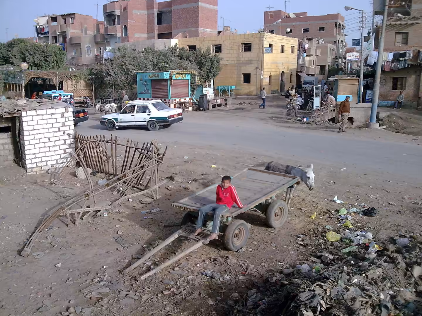 Donkey pulling wooden cart with child sitting on it in dusty residential street with apartments and small shops.