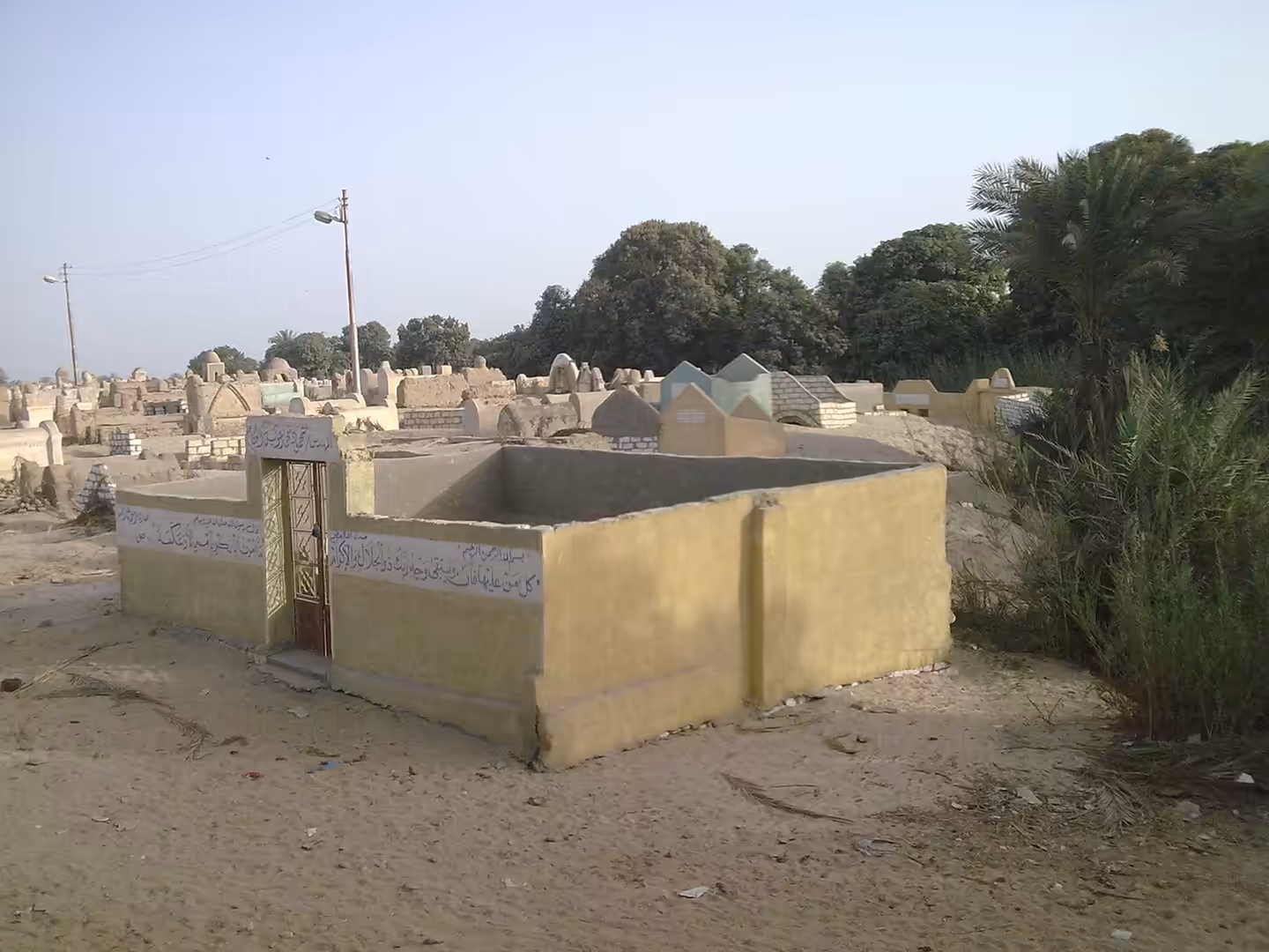 Sand colored cemetery tomb structures with Arabic writing surrounded by trees and palms.