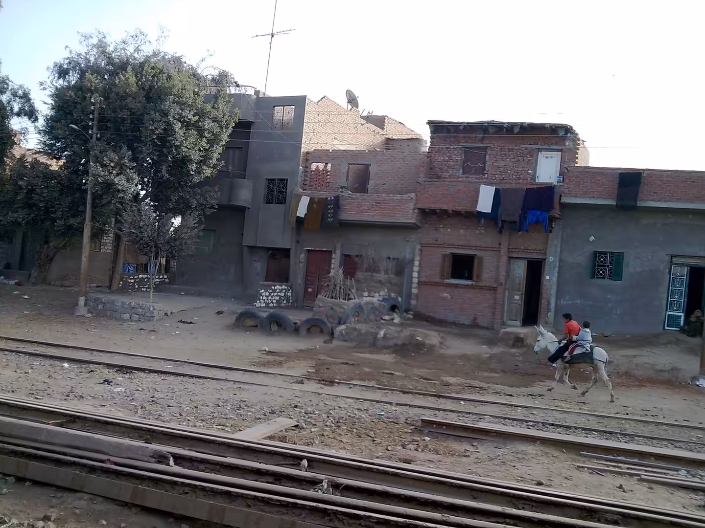 Child riding donkey beside railway tracks in front of brick village houses.