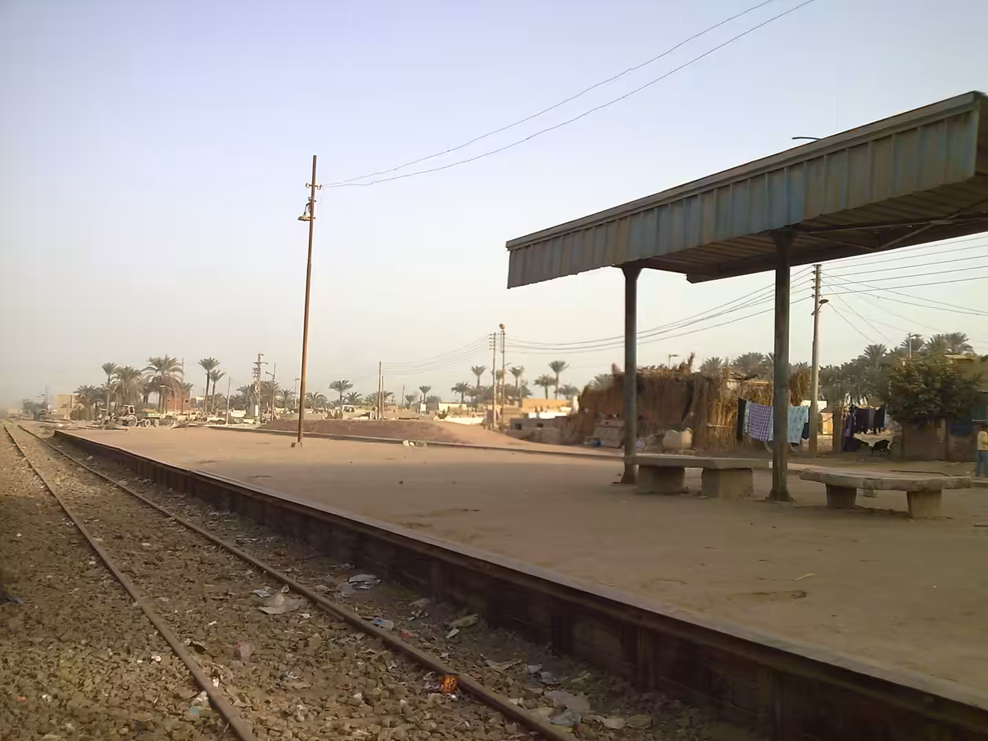 Rural train platform with metal shelter, benches, palm trees, and surrounding village landscape.