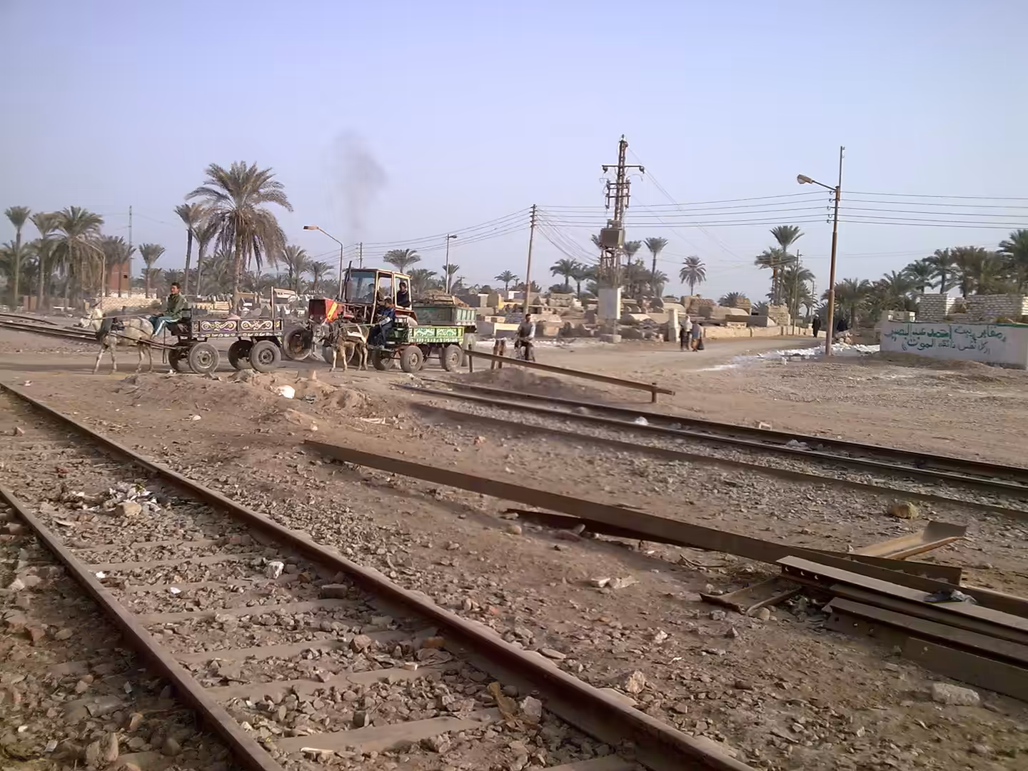 Donkey carts crossing railway tracks near palm trees and cemetery structures.