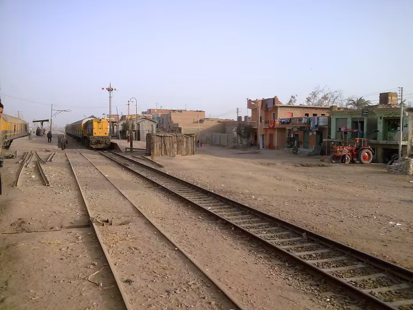 Diesel locomotive stopped at rural station with village houses and tractor nearby.