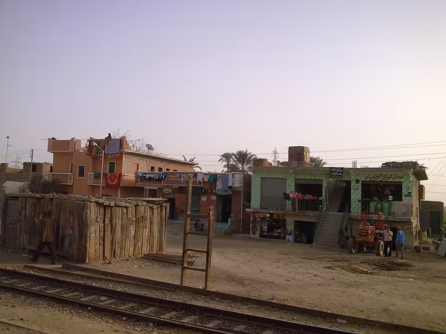 Rail tracks beside small village shops with Arabic signs, laundry hanging, and tractor parked outside.