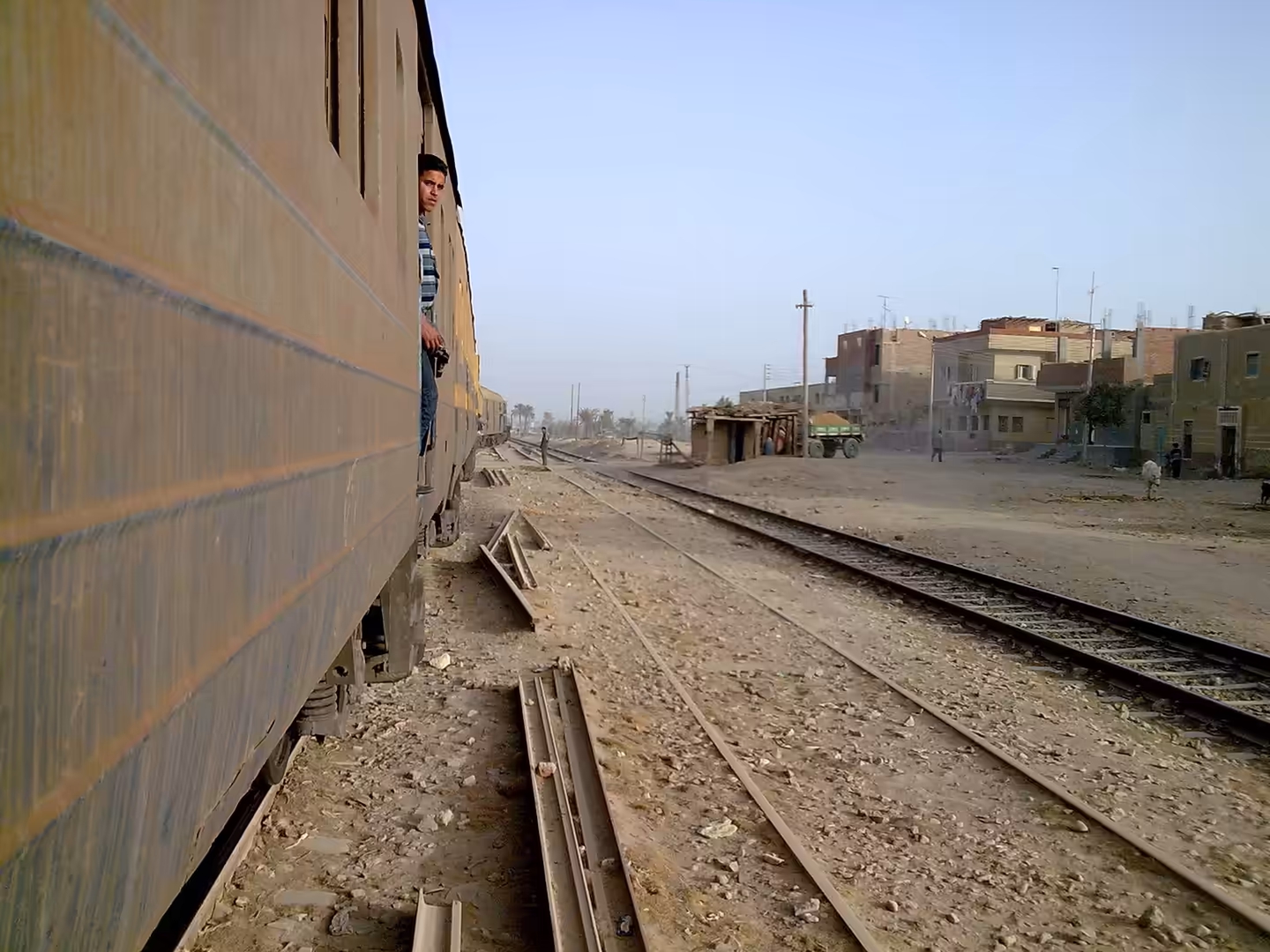 View from passenger train passing small rural station with village houses and tractor.