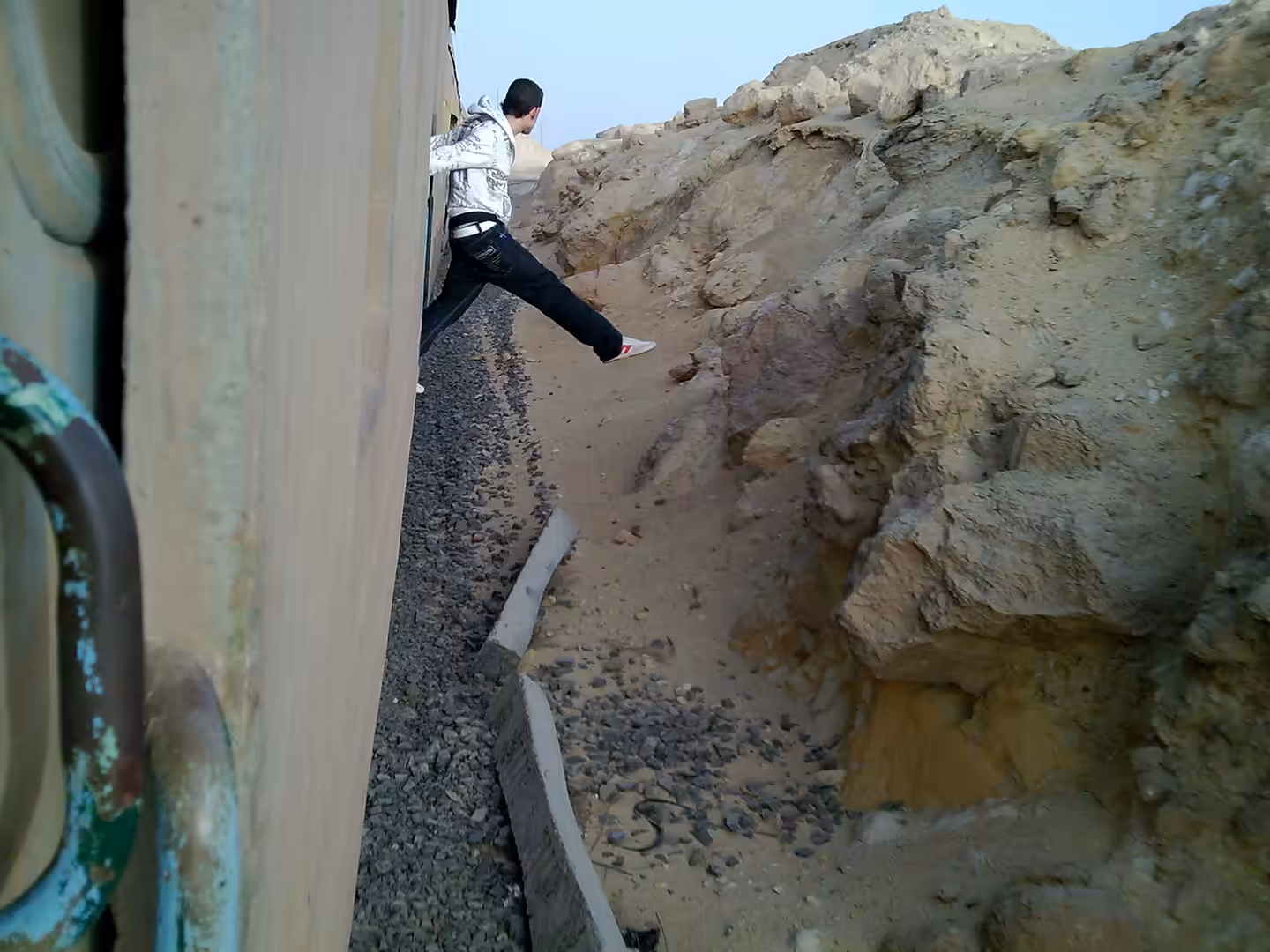 Passenger leaning from moving train beside rocky desert embankment and gravel track.