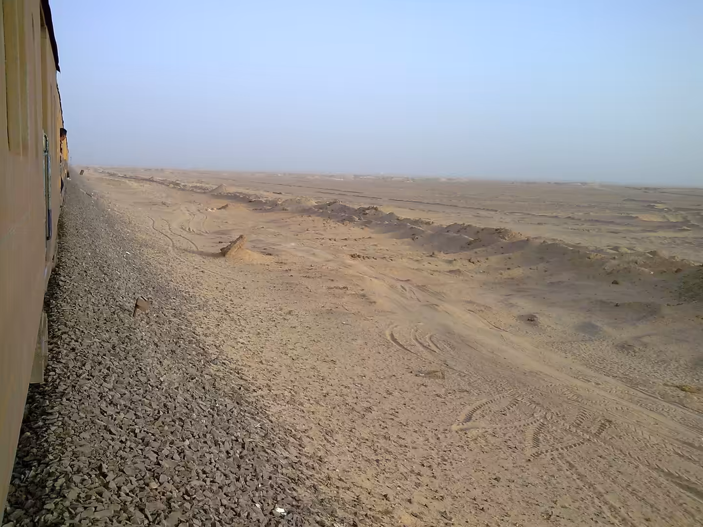 View from train along ballast track through open sandy desert with tire marks and low dunes.
