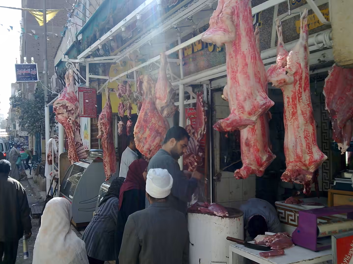 Butcher cutting meat in street shop with raw carcasses hanging outside and customers nearby.