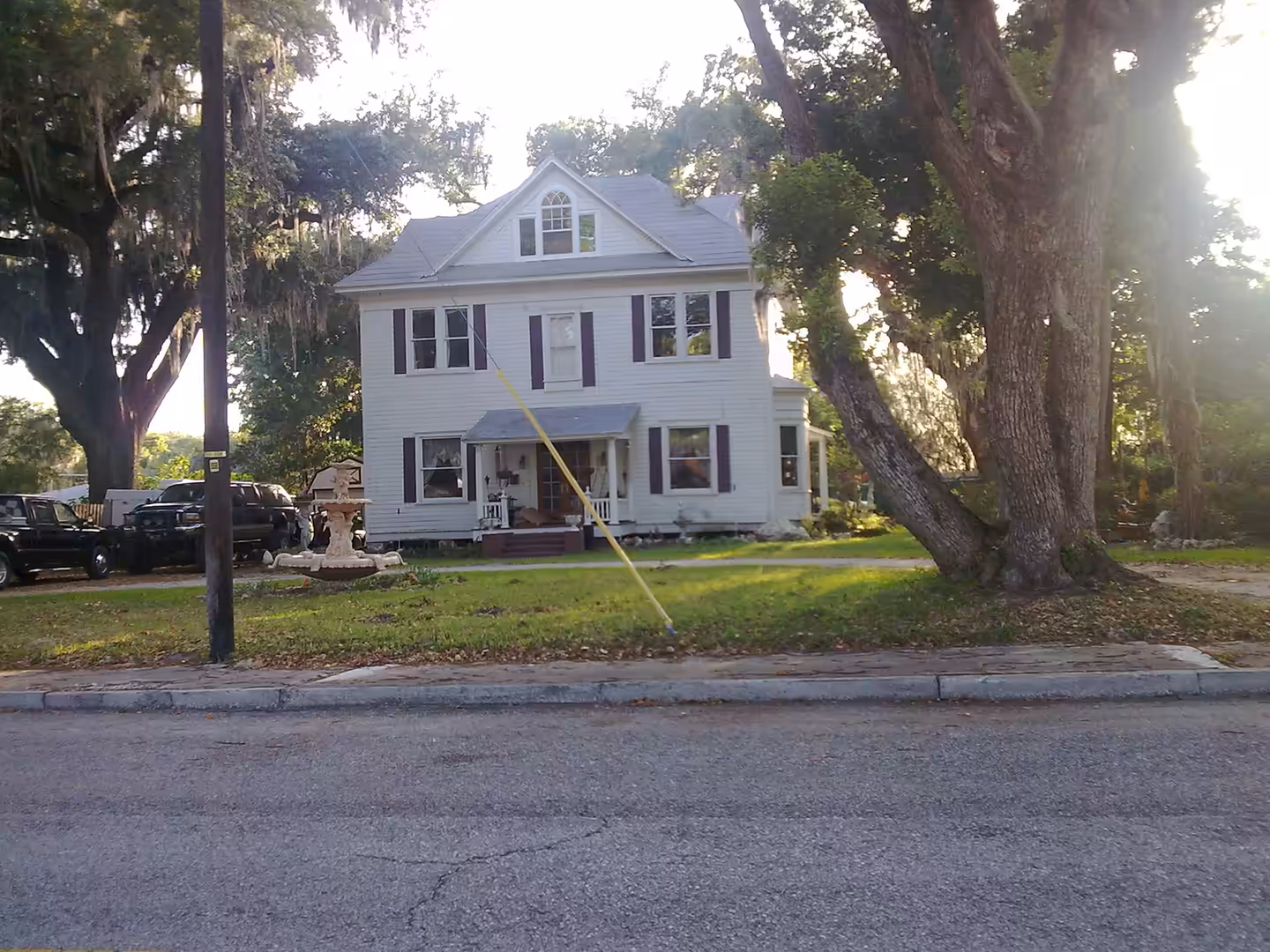 A white two-story wooden house set back from the road, framed by large oak trees and afternoon light.