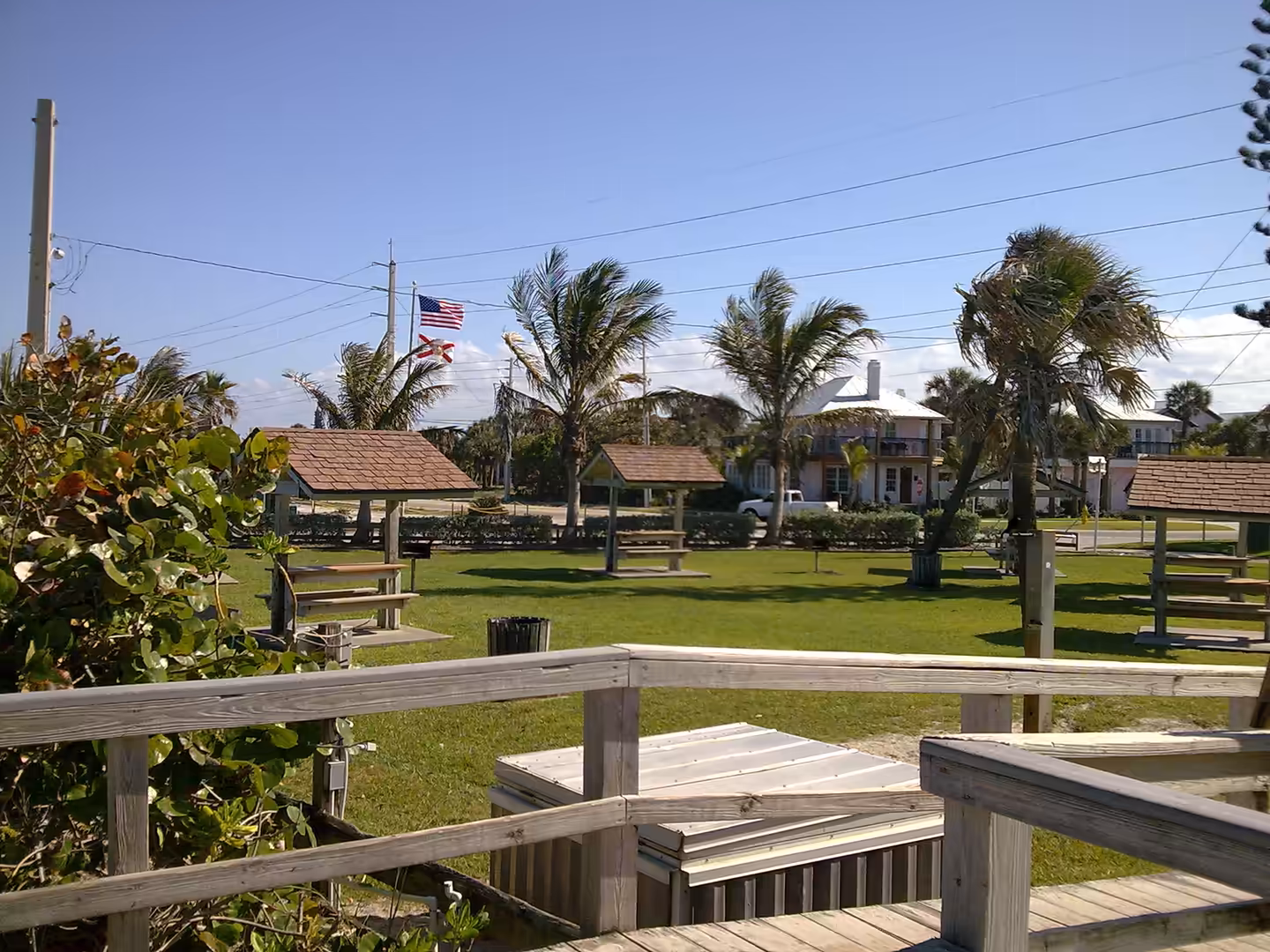 A coastal park with picnic shelters, palm trees, and flags waving near the shoreline.