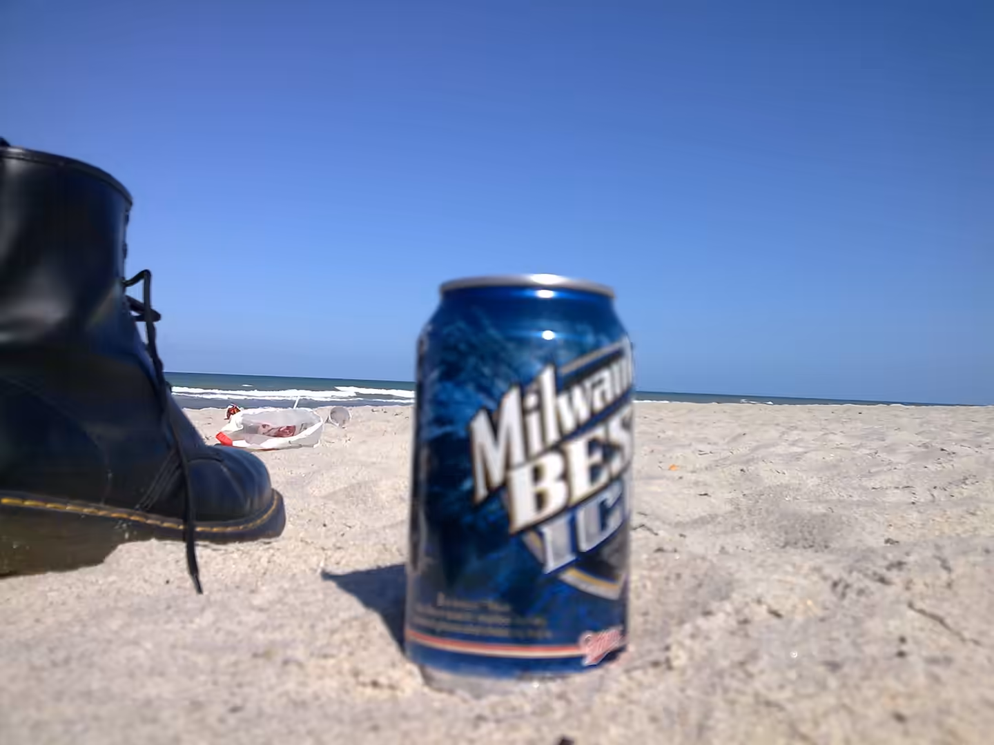 A beer can and a black boot resting on sand with the ocean and horizon in the background.