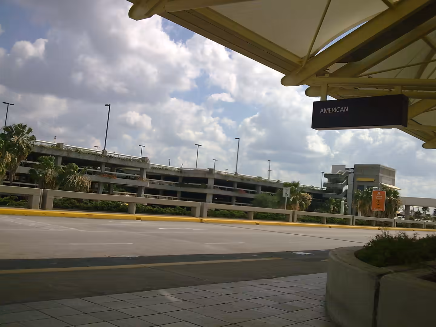 Departure curb at Orlando International Airport with covered walkway, roadway, parking garages, and palm trees under a cloudy sky.