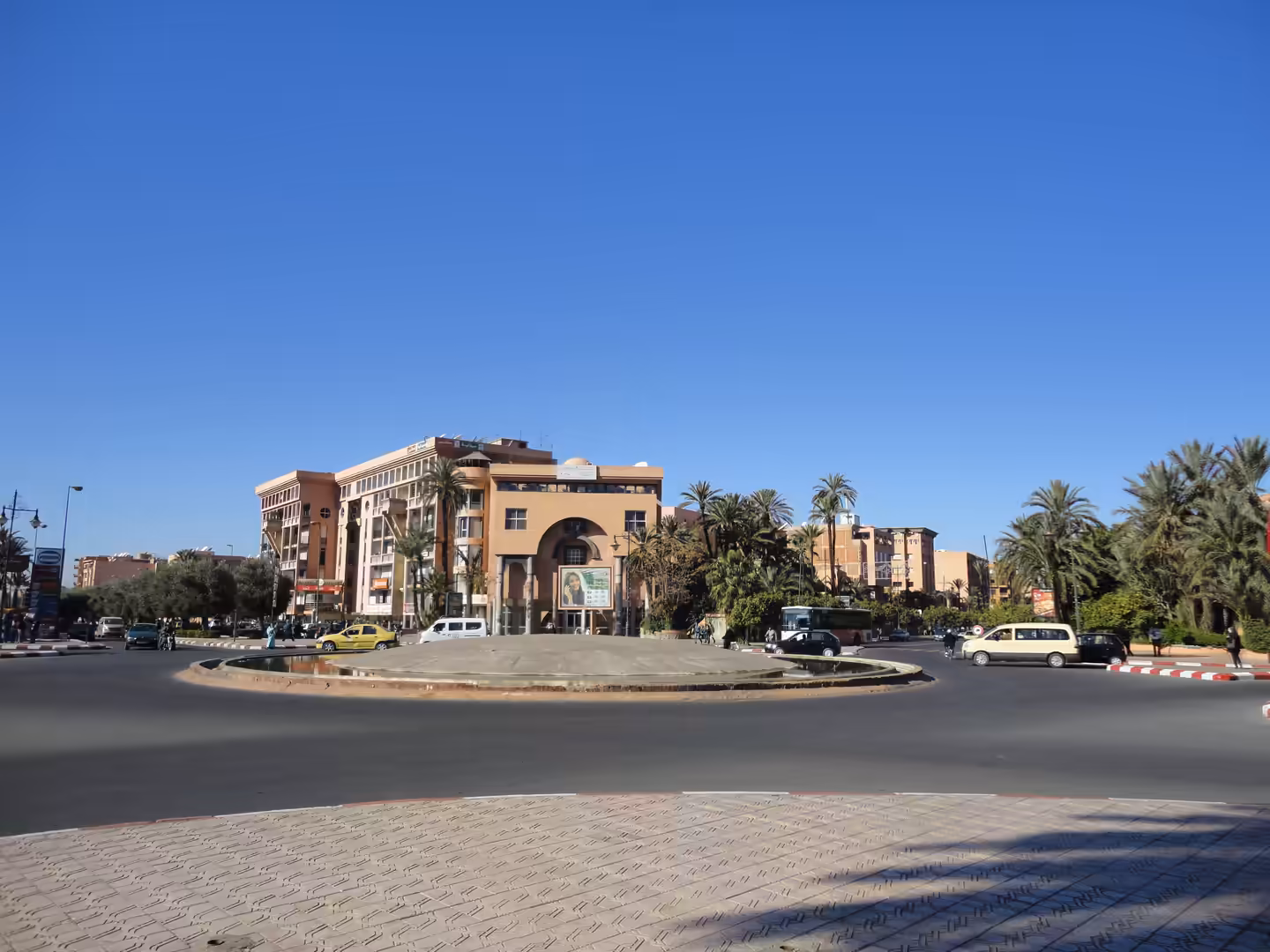 Wide urban roundabout with cars, palm trees, and terracotta-colored buildings beneath a clear blue sky.