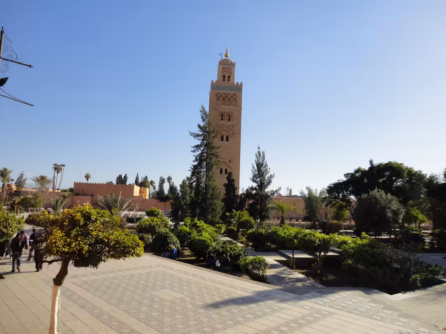 The Koutoubia Mosque minaret in Marrakech rising above green gardens and palm trees under a clear blue sky.