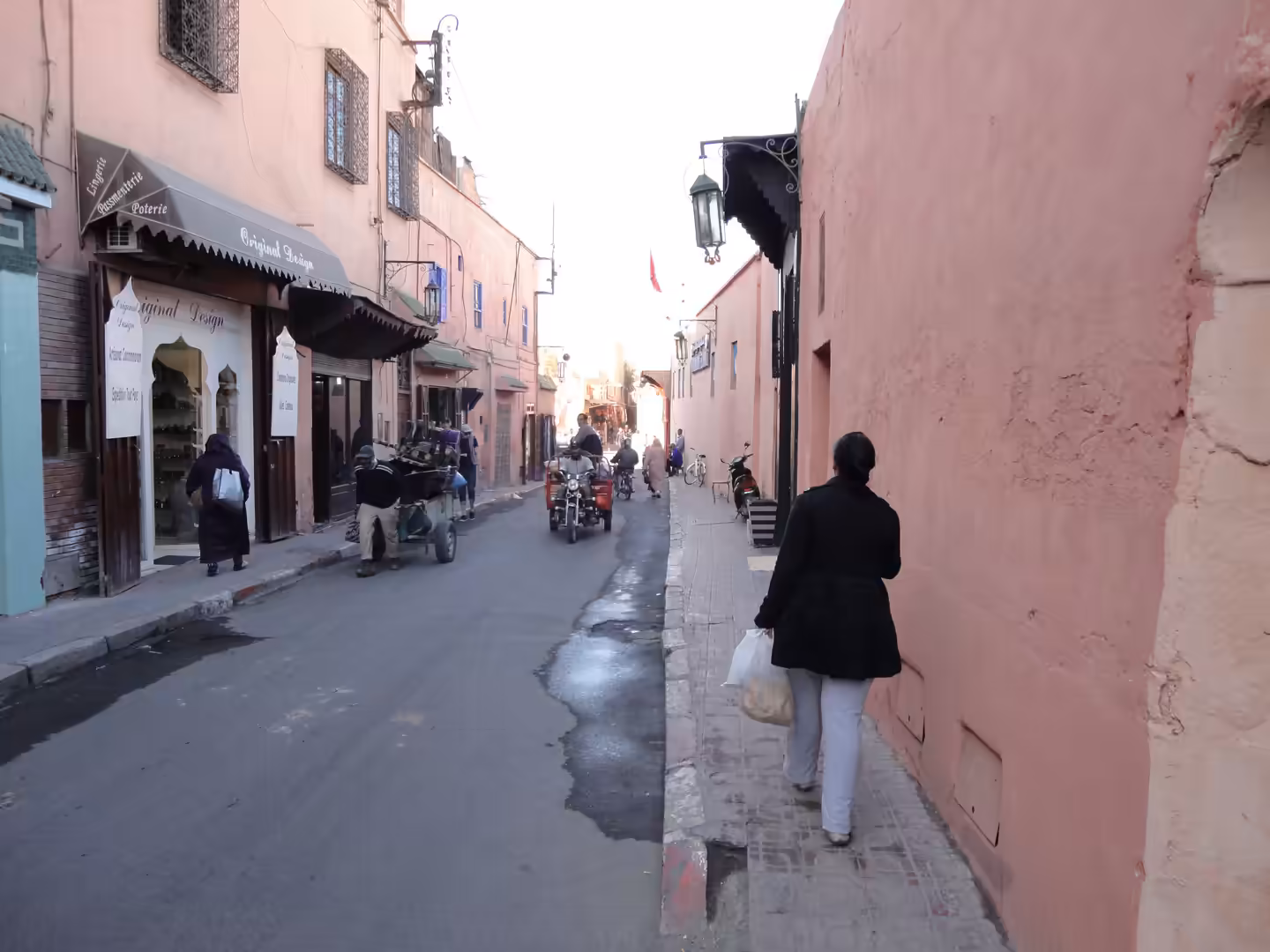 A narrow street in the Marrakech medina with pink plaster walls, pedestrians walking, and scooters driving past small shops.
