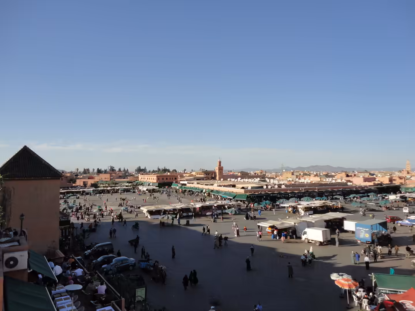 A wide aerial-style view of Jemaa el-Fnaa in Marrakech with crowds, market stalls, vehicles, and surrounding buildings under a clear sky.