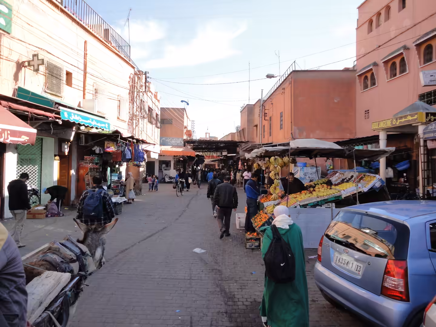A busy market street in Marrakech with pedestrians walking past fruit stalls, small shops, parked cars, and vendors under soft afternoon light.