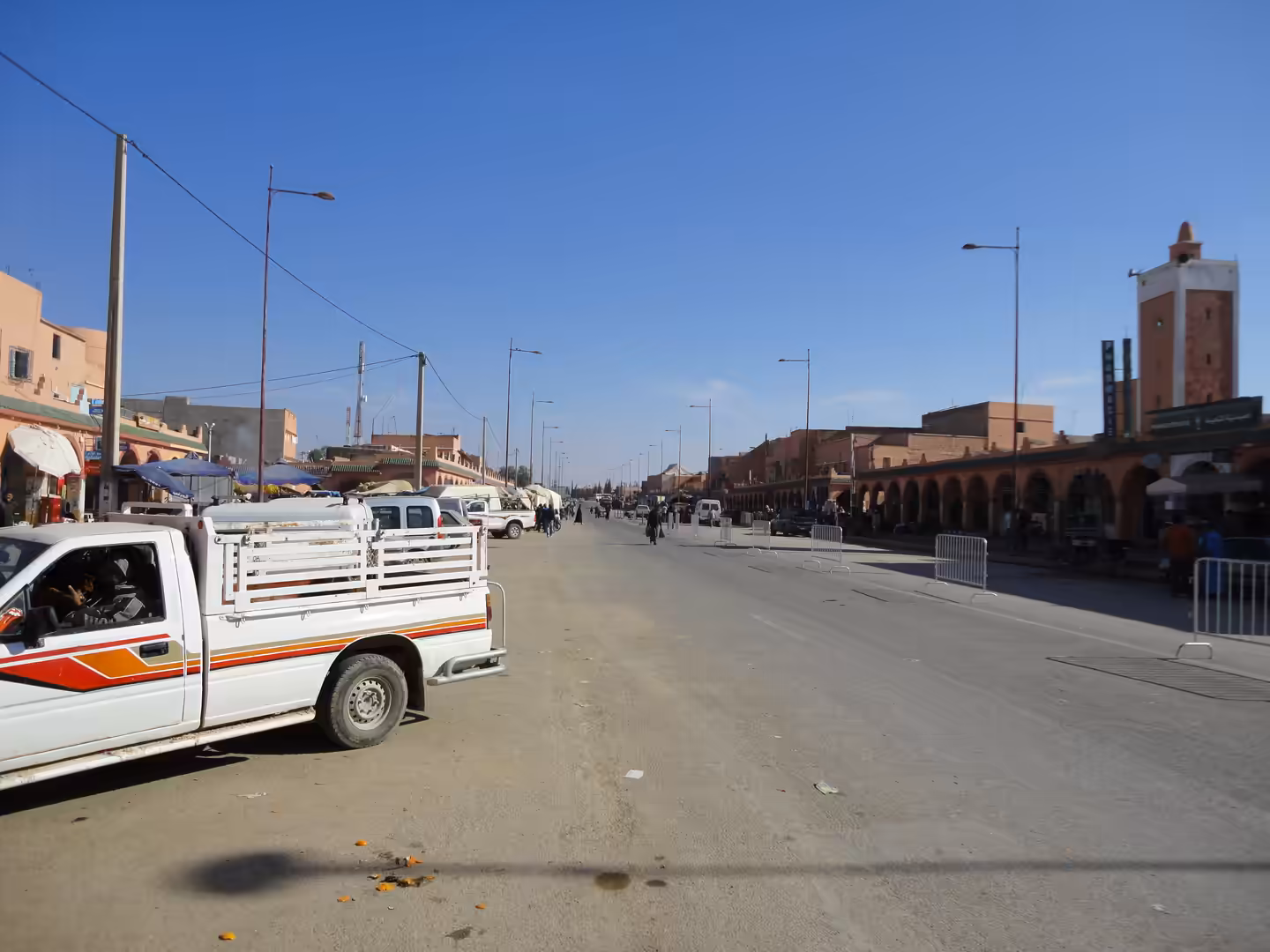 Wide street in Essaouira with a white pickup truck in the foreground, market stalls on the left, arcaded buildings on the right, and people walking in the distance under a clear blue sky.