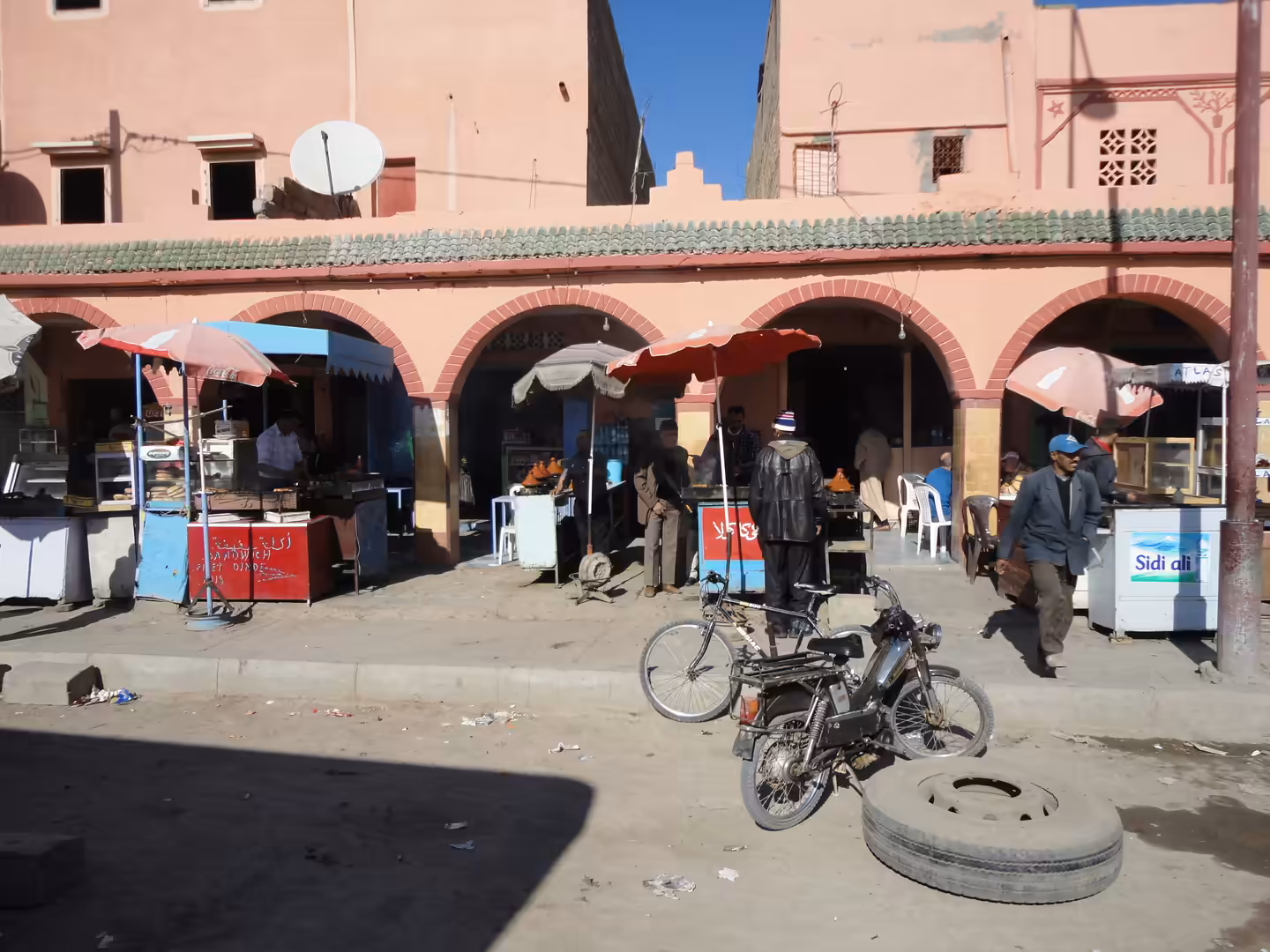 Street scene in Essaouira with arched arcades, small food stalls, men standing and talking, and a bicycle parked beside a loose tire on the road.