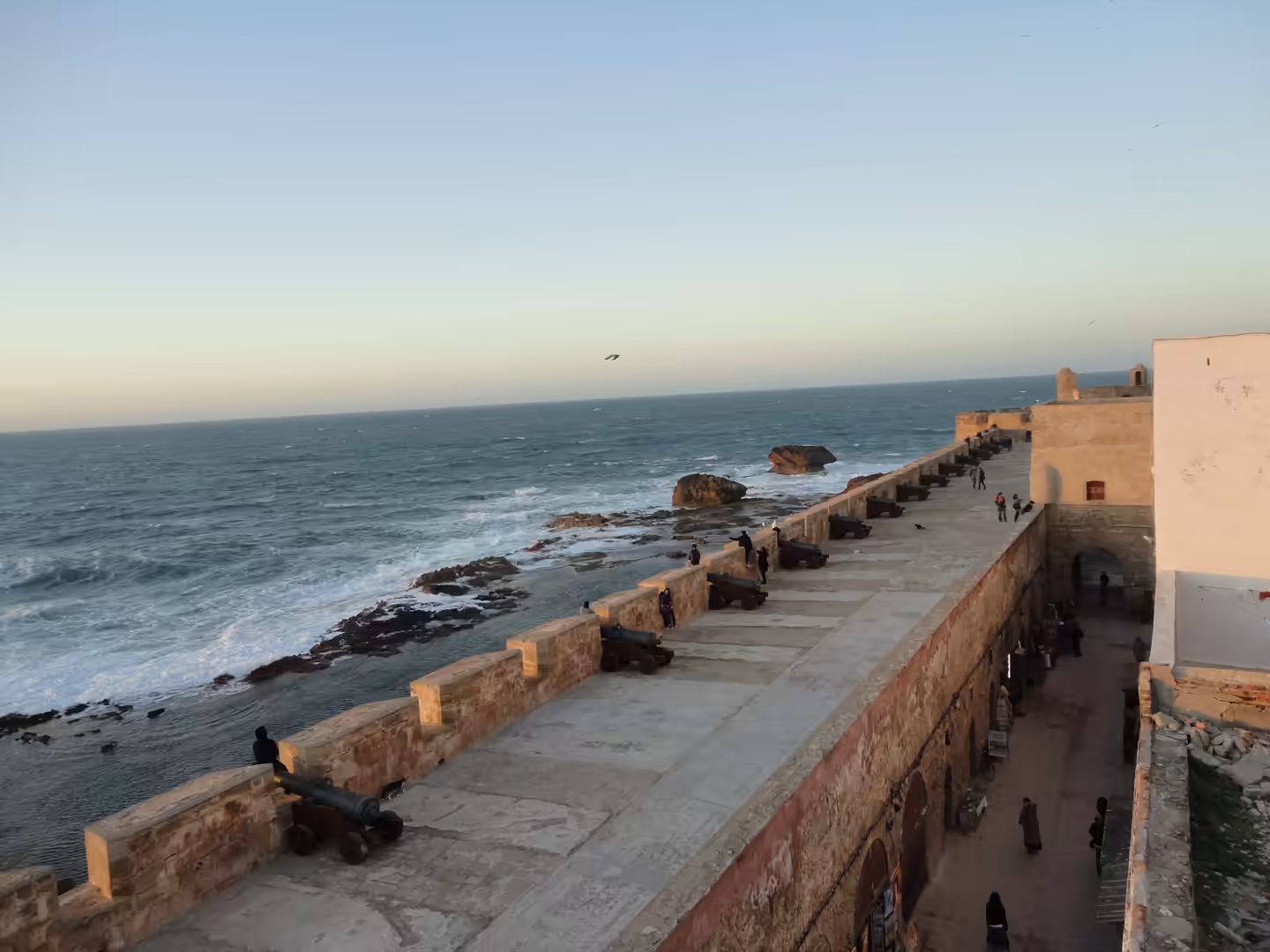 Stone ramparts in Essaouira with old cannons facing the Atlantic Ocean, waves breaking against rocks on the left, and people walking along a lower passage on the right.