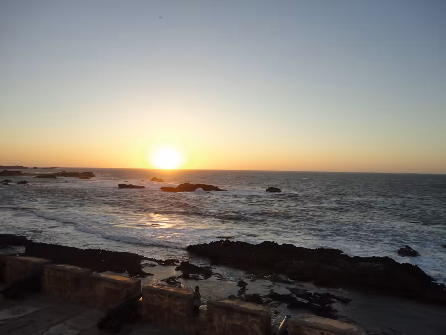 Sun setting over the Atlantic Ocean with waves rolling toward rocky outcrops, seen from stone ramparts in Essaouira.