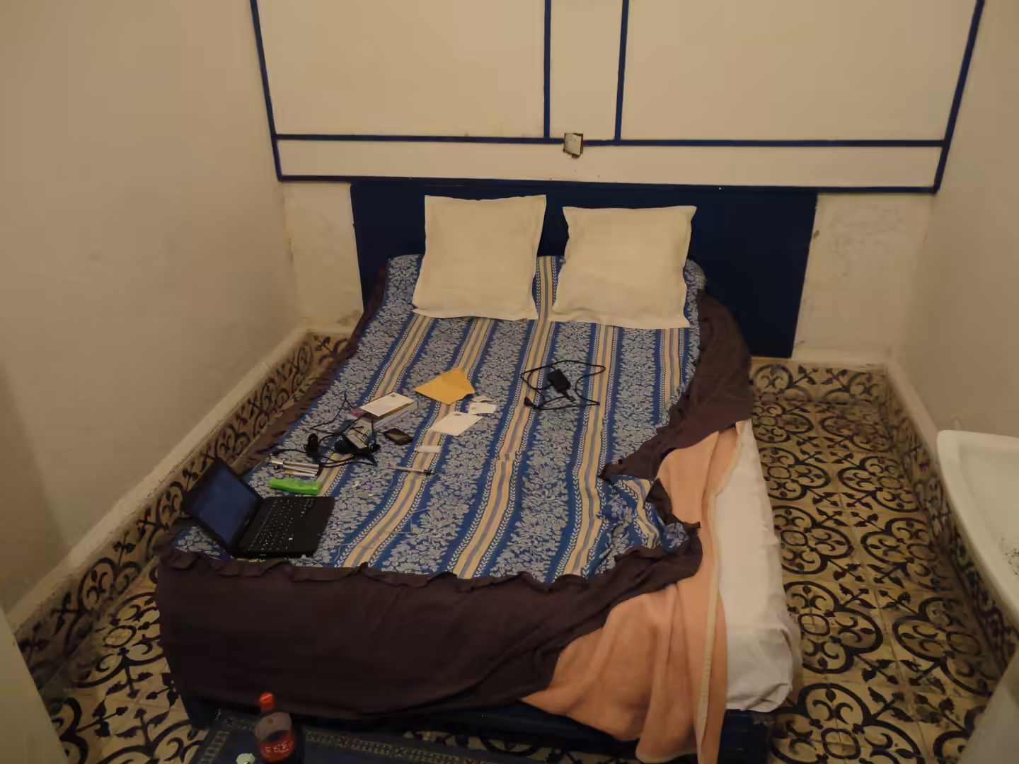 A double bed with striped bedding and two pillows in a small room, with a laptop, cables, papers, and personal items placed on the bed.