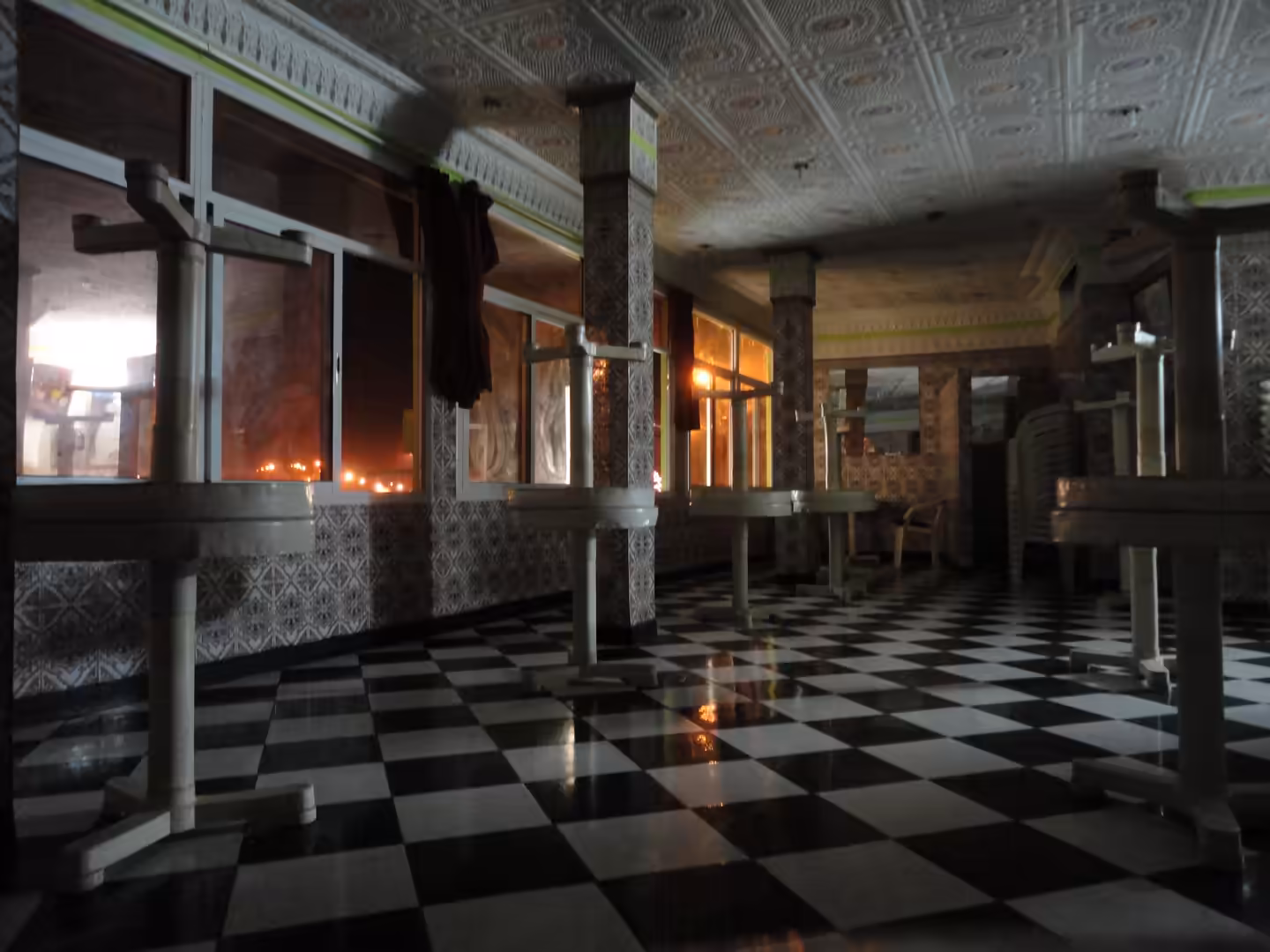Empty interior hall at night with patterned tiled columns, black-and-white checkered floor, metal tables, and windows lit by orange streetlights outside.
