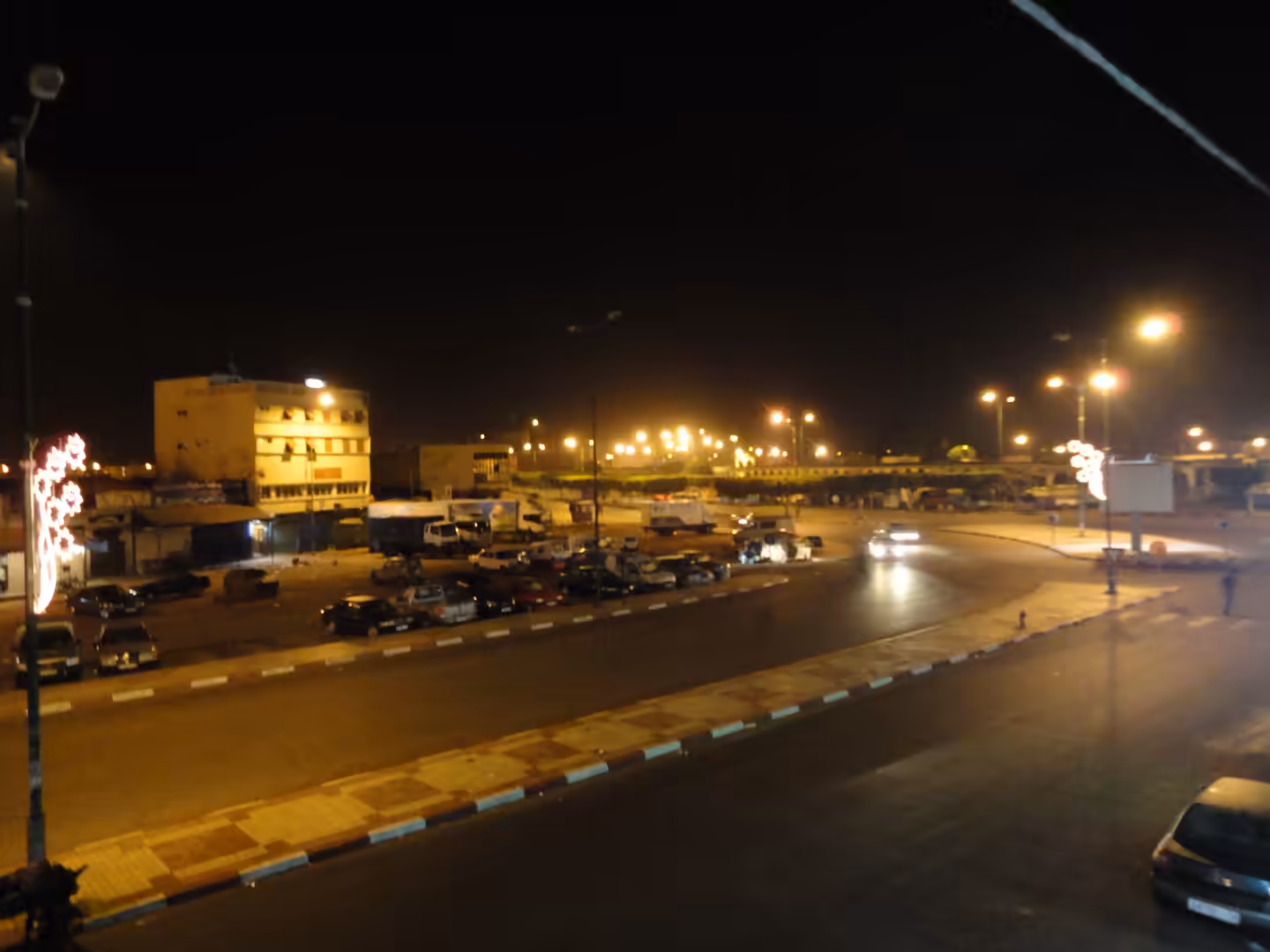 Night view of a wide intersection in Inezgane with cars driving through, parked vehicles along the curb, streetlights, and low-rise buildings lit by warm yellow light.