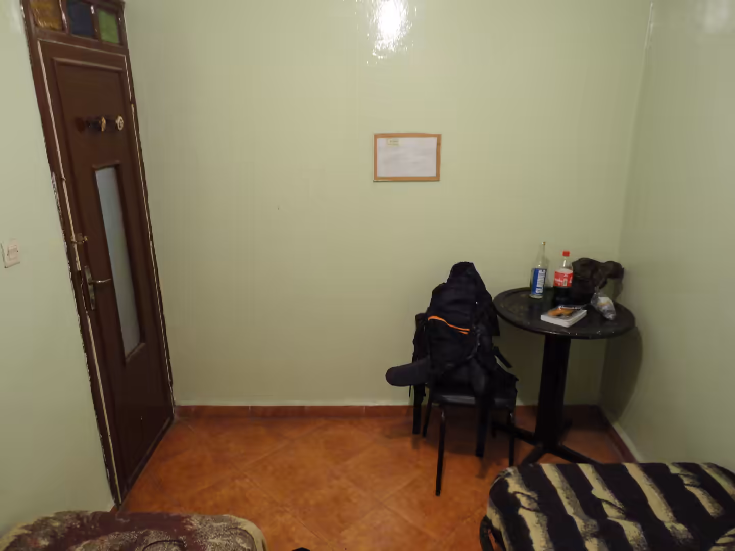 Corner of a small room with pale green walls, a chair holding a backpack, a small round table with bottles and snacks, tiled floor, and a wooden door.