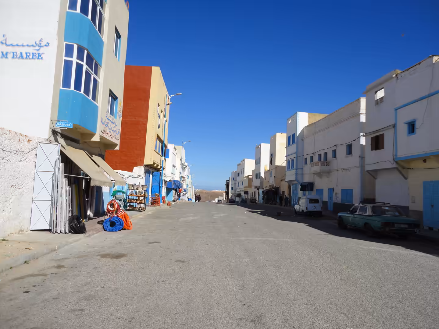 An empty street in Sidi Ifni with white and blue buildings on both sides, parked cars, small shops, and a deep blue sky overhead.