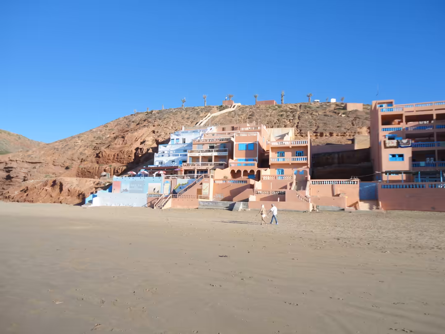 Pastel-colored houses built into a rocky hillside overlook a wide sandy beach at Legzira, with two people walking across the foreground under a clear blue sky.
