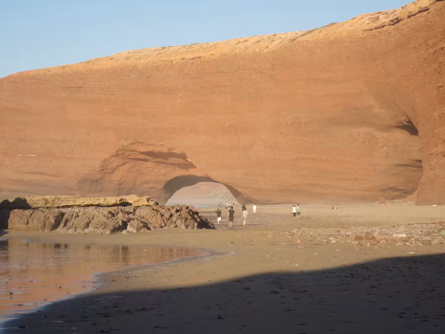 People standing on the beach beneath the still-existing red sandstone sea arch at Legzira in 2011, with the cliff face and tidal flats visible.