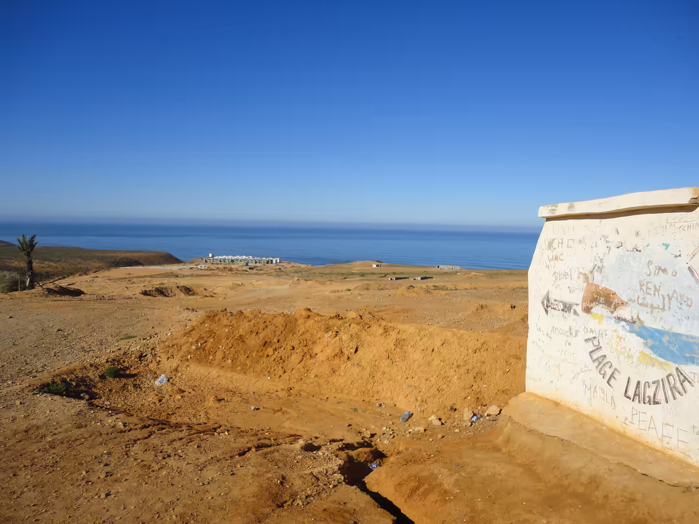 Dry cliff-top lookout above Legzira Beach with a graffiti-covered concrete marker, sandy ground, and a wide Atlantic horizon under a cloudless sky.
