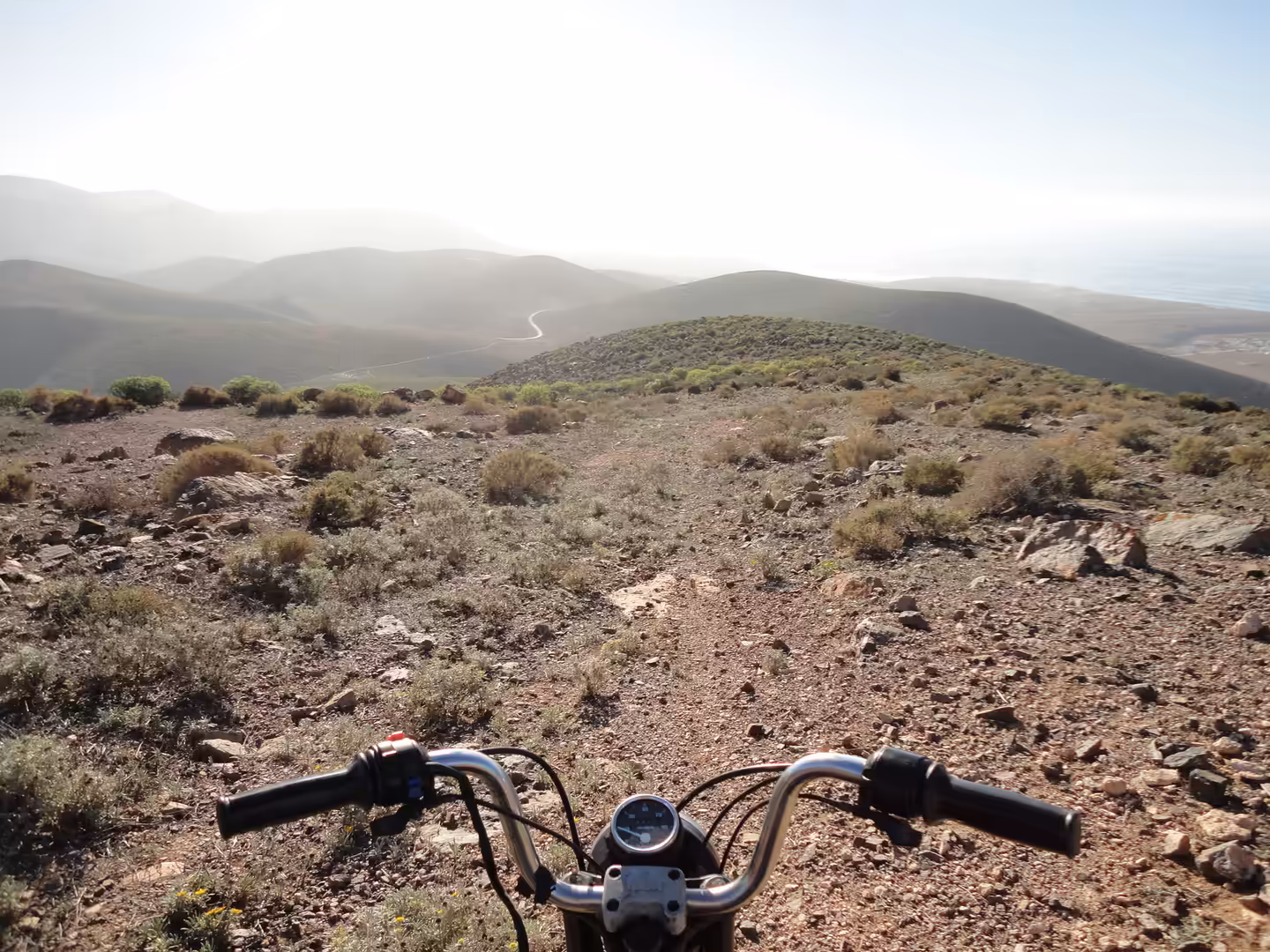 Motorcycle handlebars in the foreground overlooking rocky hills, sparse shrubs, and winding tracks across vast valleys.