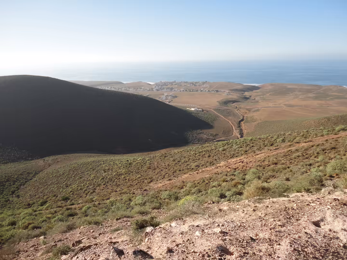 Steep hillside overlooking green-brown valleys descending toward a small coastal settlement and the Atlantic Ocean.