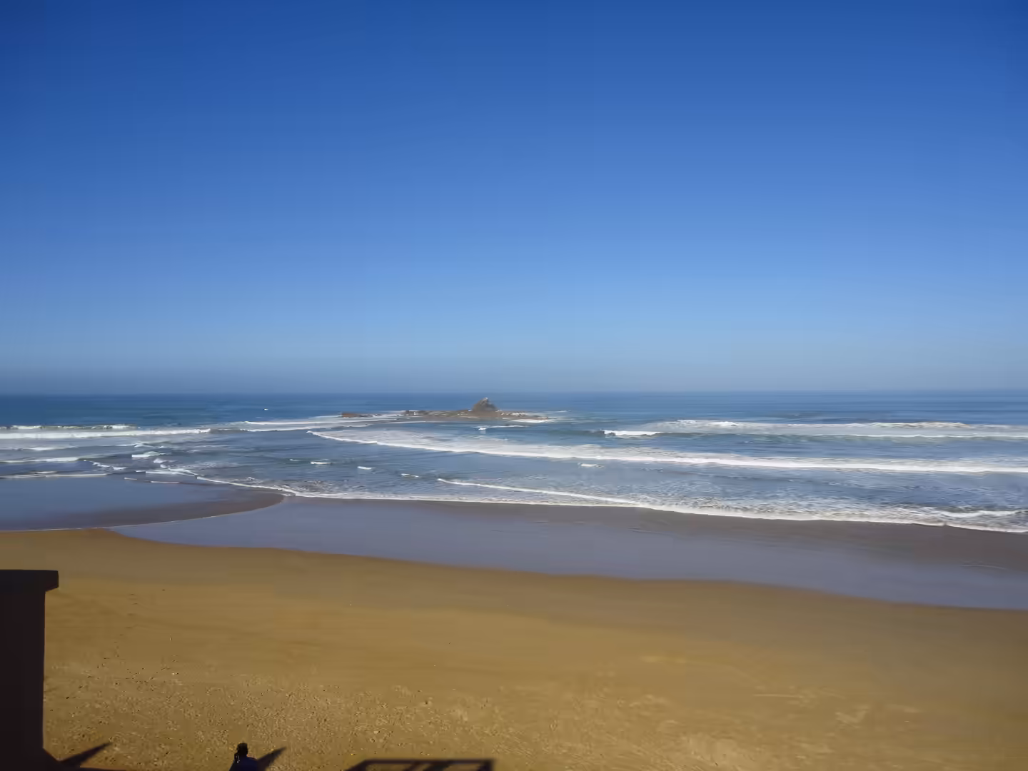 Wide sandy beach with Atlantic waves rolling in and a rocky sea stack offshore under a clear blue sky.