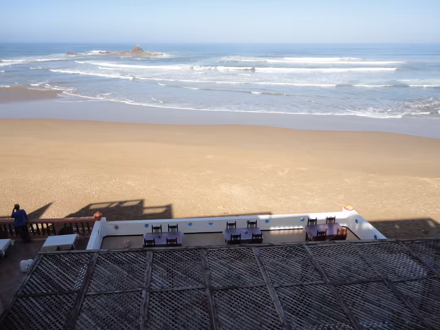 Terrace with wooden tables overlooking Legzira Beach, wide golden sand, Atlantic waves, and a rocky outcrop offshore.