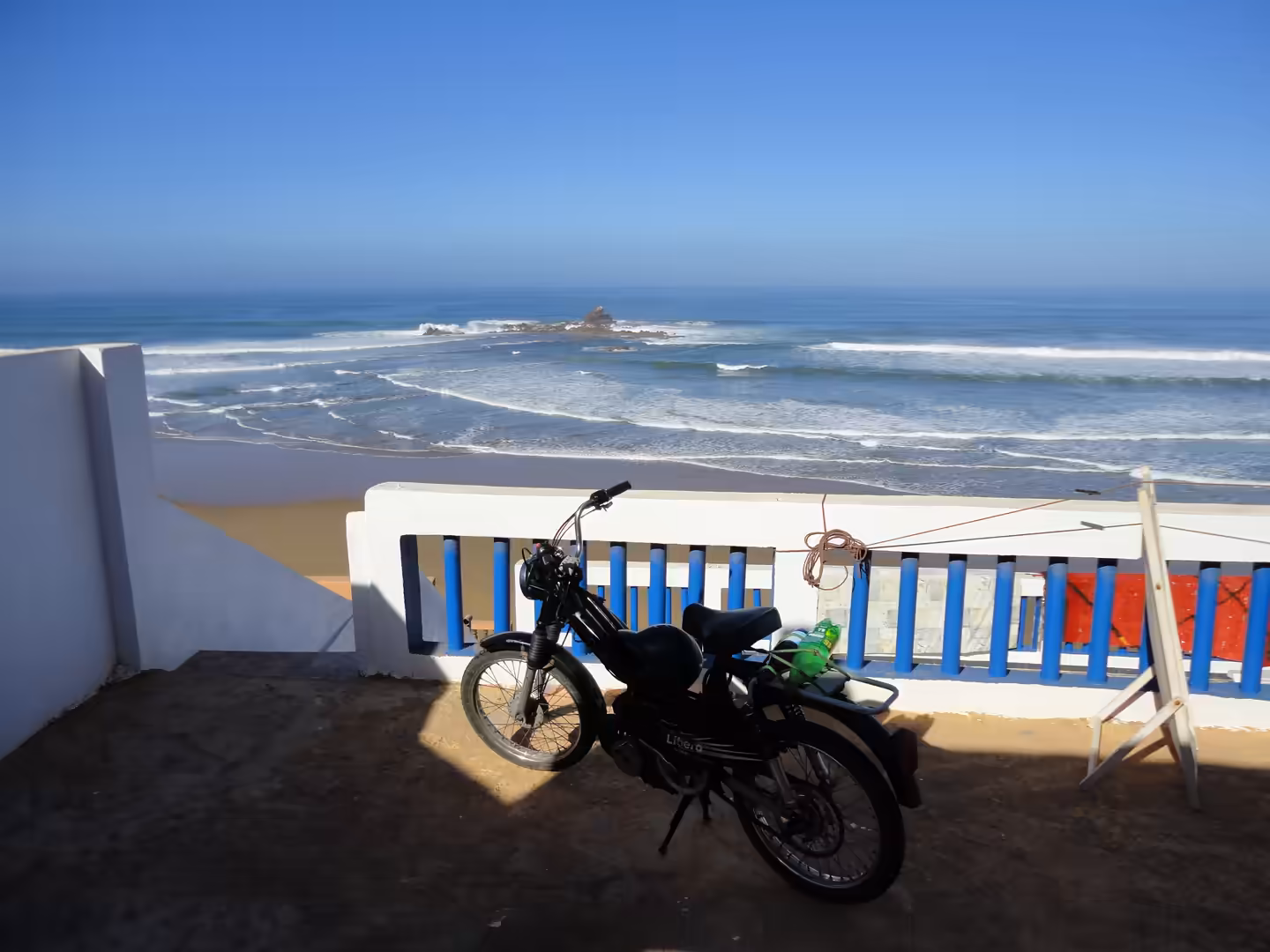 Motorcycle parked on a terrace overlooking Legzira Beach, with Atlantic waves and a rocky outcrop offshore under a clear sky.