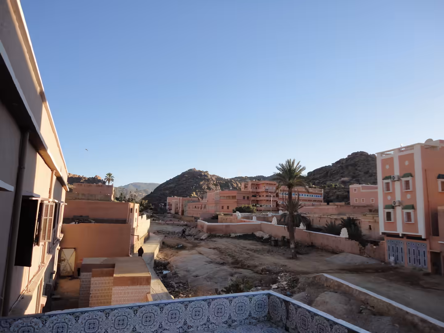 View across Tafraoute with pink-toned buildings, palm trees, rocky ground, and surrounding Anti-Atlas hills under a clear sky.