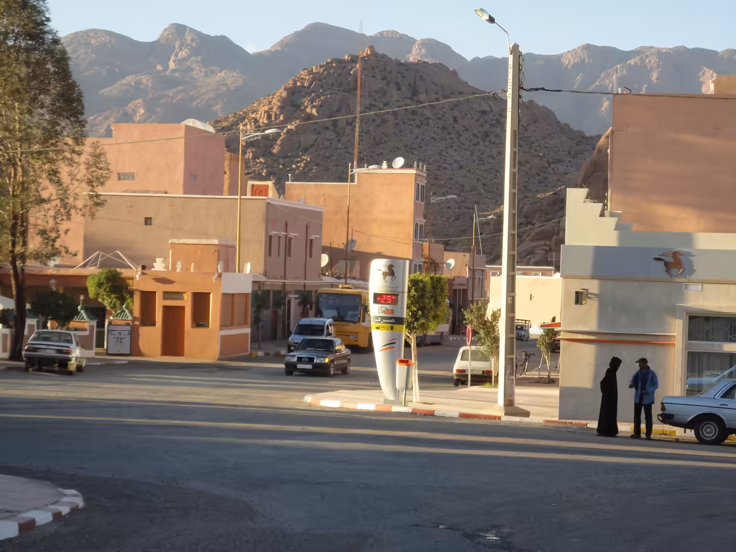 Street scene in Tafraoute with pastel buildings, cars, pedestrians, and rugged granite mountains rising behind the town.