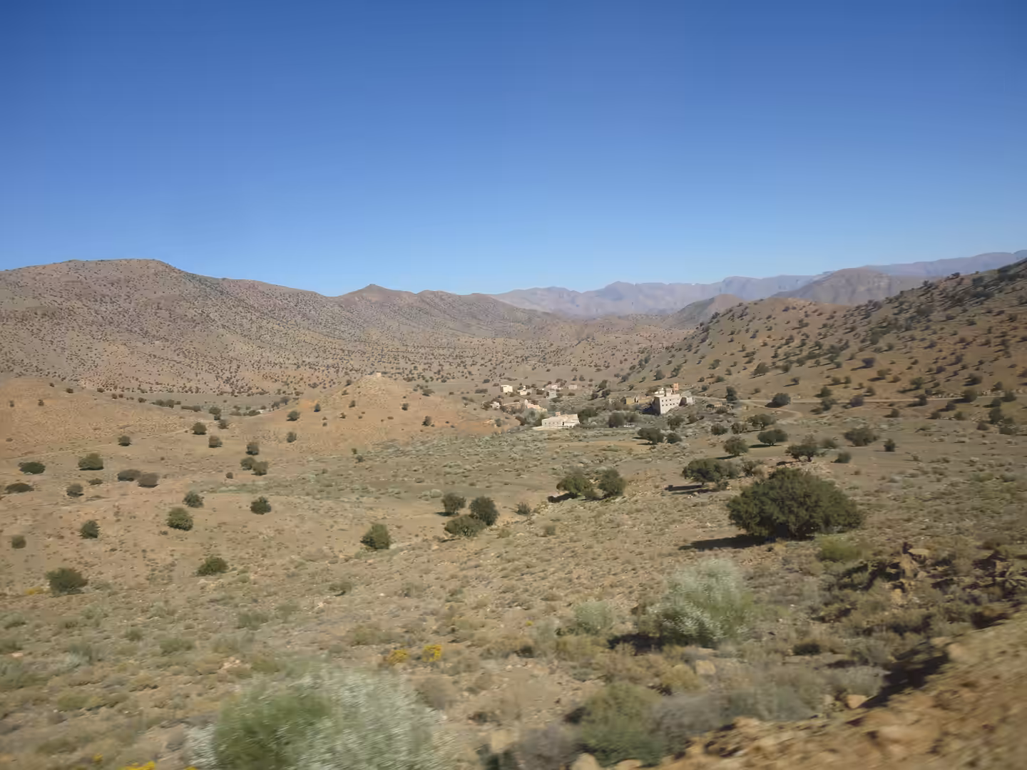 Wide arid valley with scattered shrubs, small hilltop village, and rounded Anti-Atlas mountains under a clear blue sky.