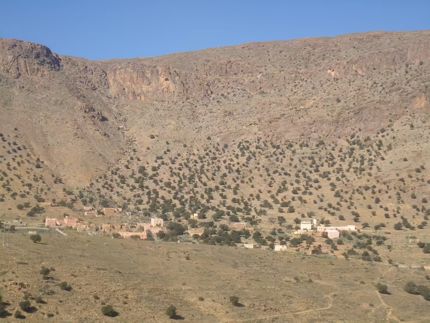 Small mountain village spread across a dry plain beneath steep, shrub-dotted cliffs in the Anti-Atlas.