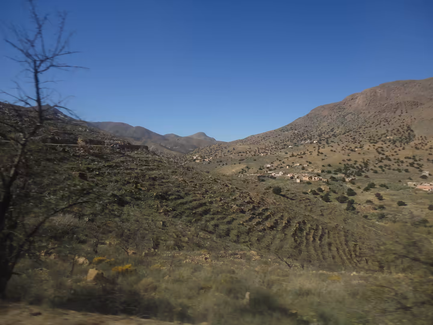 Terraced hills with sparse shrubs and small houses in a wide mountain valley under a clear blue sky.